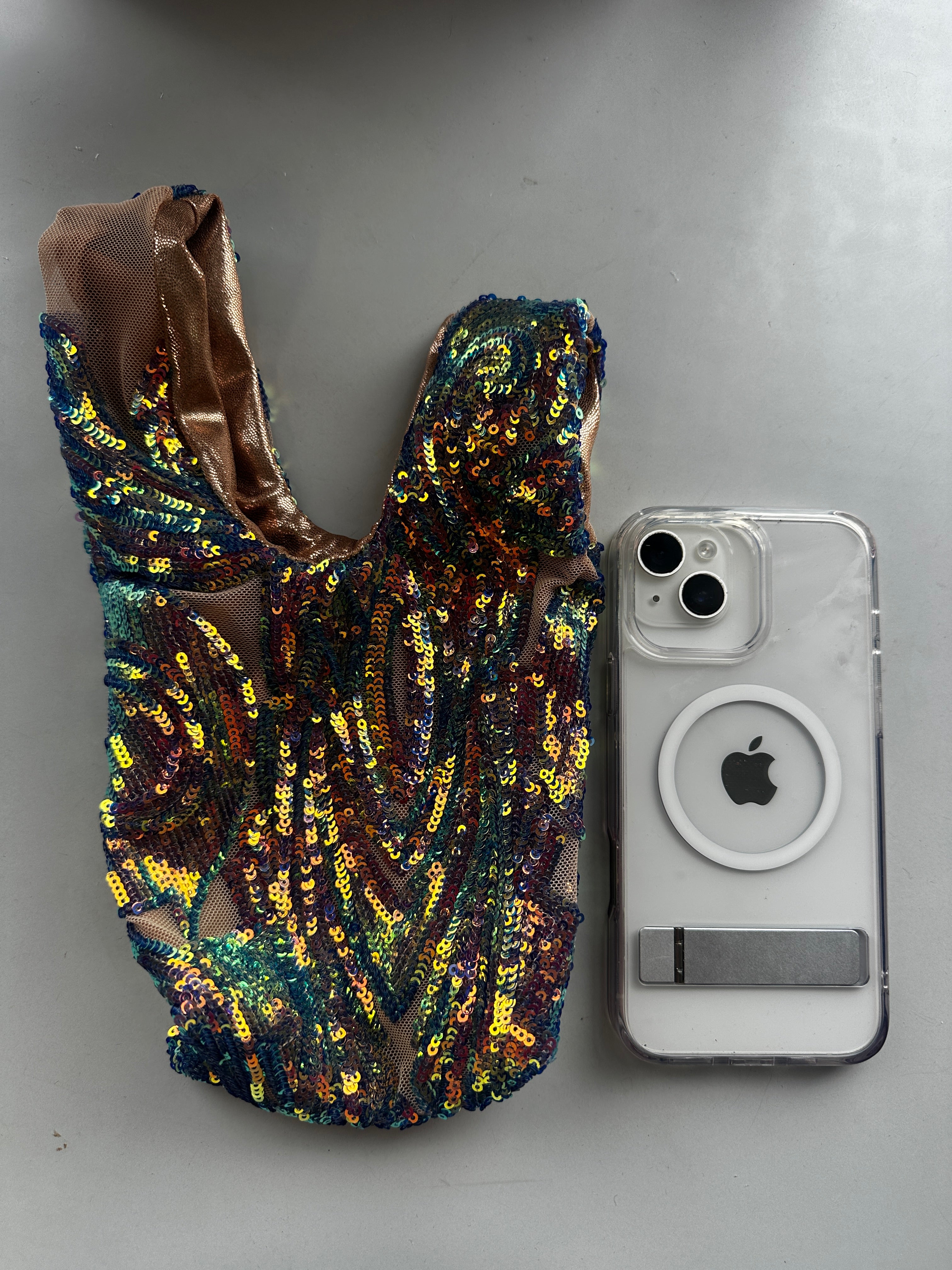 A handcrafted, reversible sequin mini tote—a sustainable fashion accessory—rests beside a clear iPhone with a MagSafe ring on a gray surface.