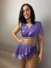 A woman poses slightly smiling in a Lavender Purple Sequin Dance Costume—featuring high waist shorts with a half skirt, crop top, and 4-way stretch mesh—against a plain light background.