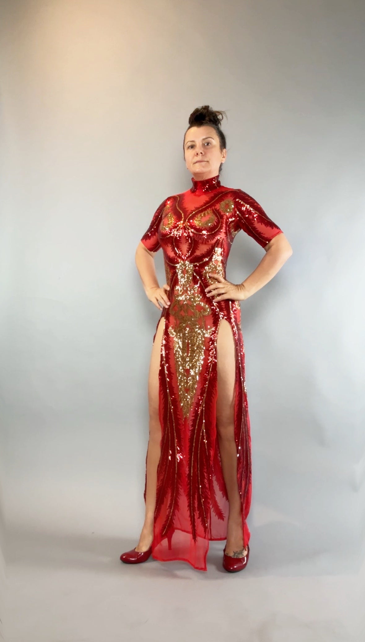 Woman wearing the Red And Gold Sequin Sheer Dress, sleeveless, with hand accessories, posing with hands on hips against a gray background.