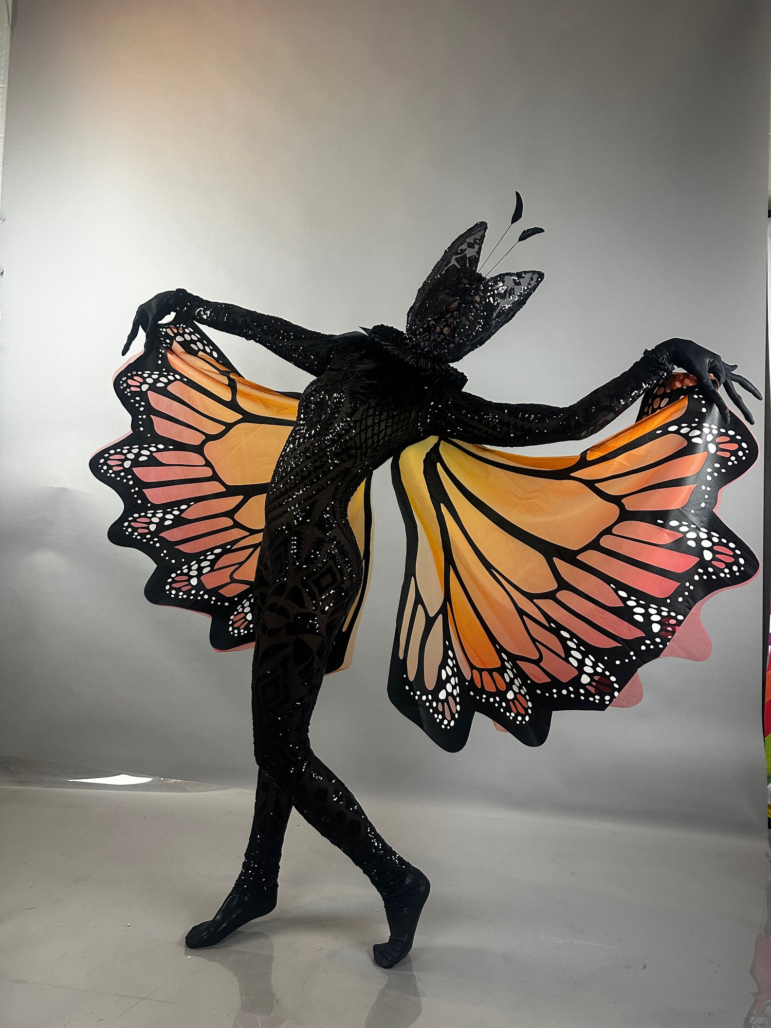 Person in a black bodysuit and orange-black butterfly wings poses dramatically against a gray backdrop, completing the look with the Black Sequin Butterfly Headpiece—a sheer mesh fairy hat with feather antennae for a festive, theatrical touch.