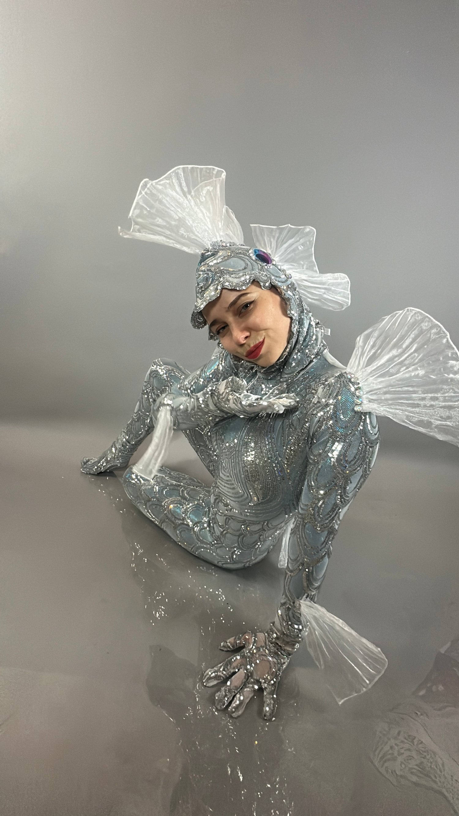 Wearing the Sea Creature costume—a shiny, silver design with clear fin details—a person sits on a reflective gray floor, showcasing exquisite couture costume craftsmanship.