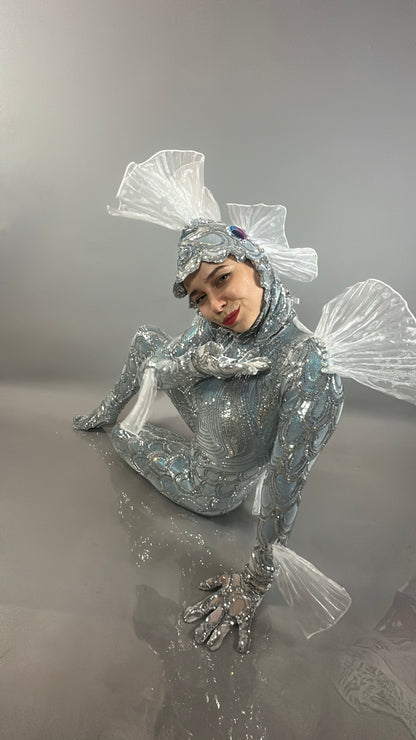 Wearing the Sea Creature costume—a shiny, silver design with clear fin details—a person sits on a reflective gray floor, showcasing exquisite couture costume craftsmanship.