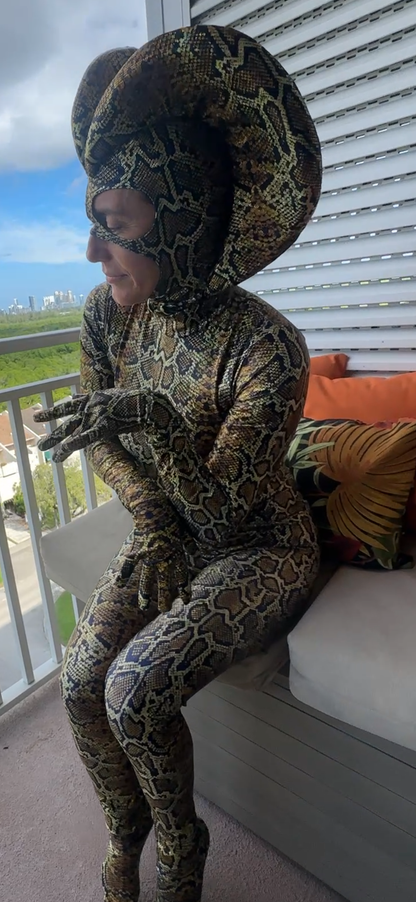Wearing a Green Cobra Snake Costume, a person in a custom acrobatics and contortionist outfit sits on a balcony couch with tropical pillows.