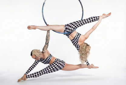 Two women wearing Asymmetrical Harlequin Catsuits—checkerboard spandex jumpsuits with one sleeve and one leg cutouts—perform an acrobatic pose with an aerial hoop against a white background.