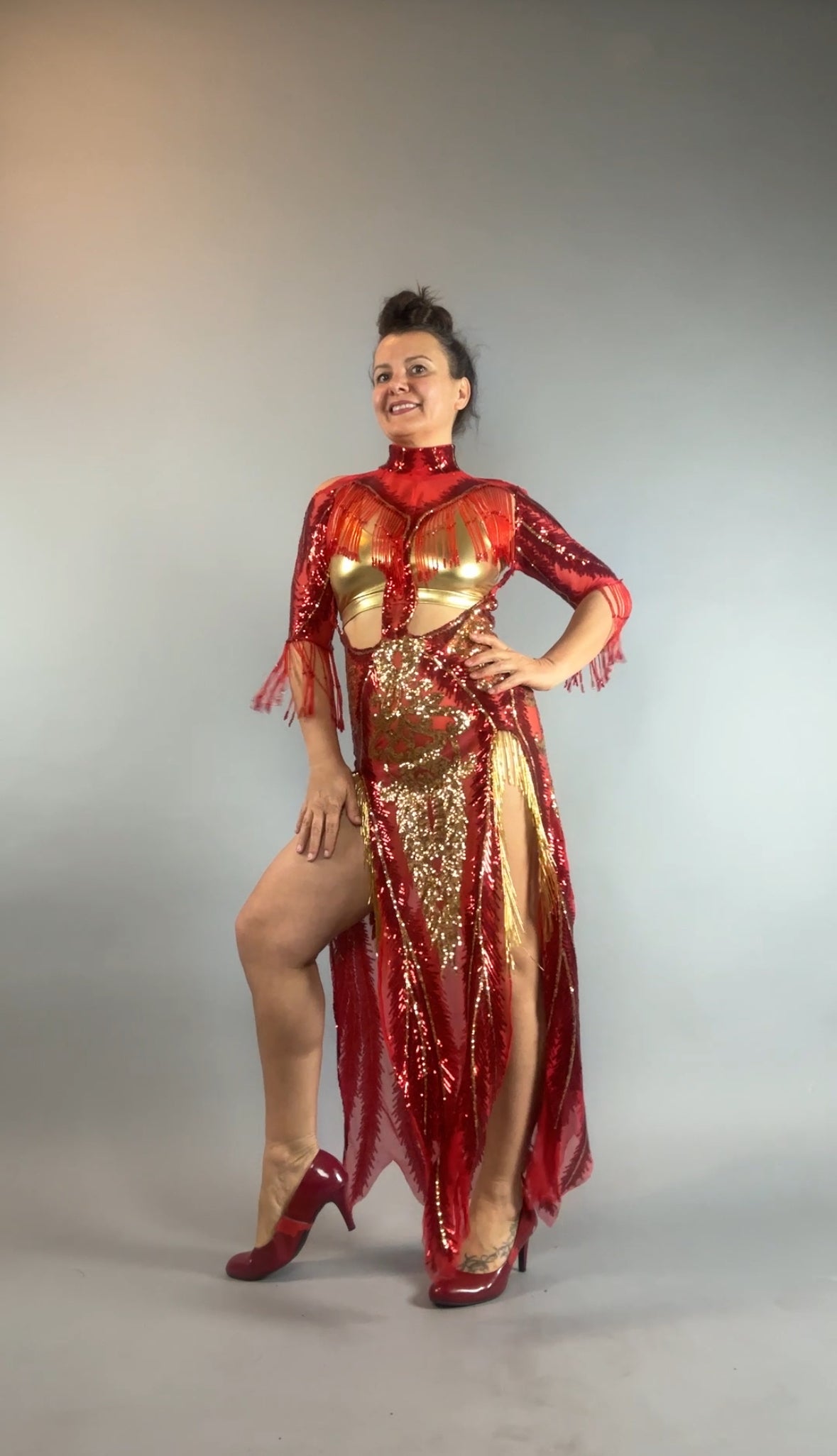 Woman poses confidently in the bearded French accessories Sequin Sheer Dress—backless with a thigh-high slit and beaded fringe accents—paired with red heels against a plain background.