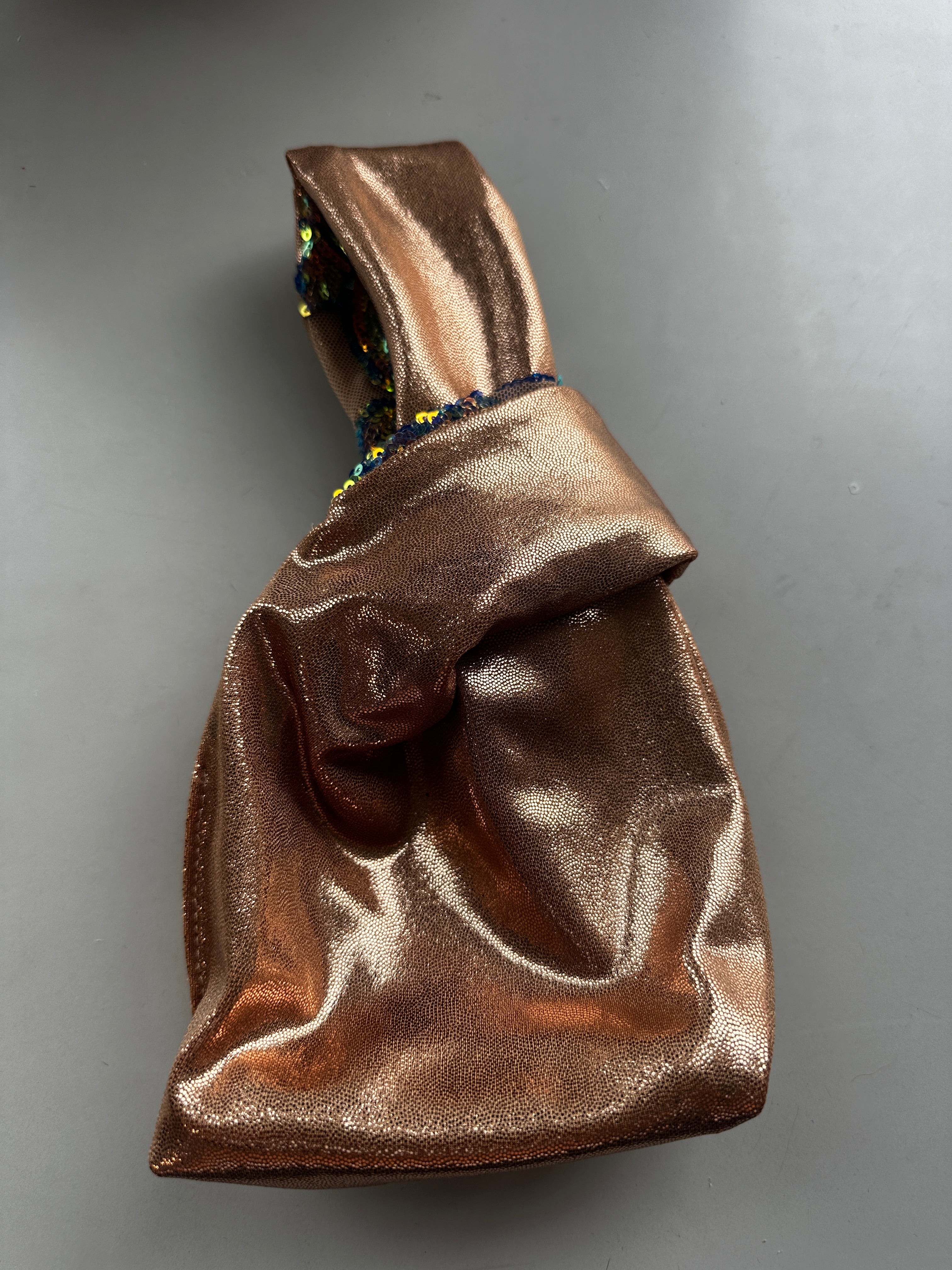 A handcrafted, reversible sequin mini tote—an upcycled sustainable fashion accessory in metallic bronze with a knotted handle—rests on a dark gray surface.