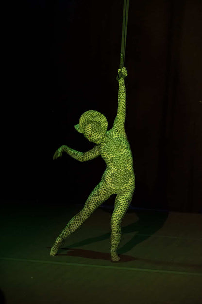 A person poses confidently on stage in the Green pixel Cobra Costume—snake print catsuit, headdress, and dance unitard—holding a strap above their head against a dark background.