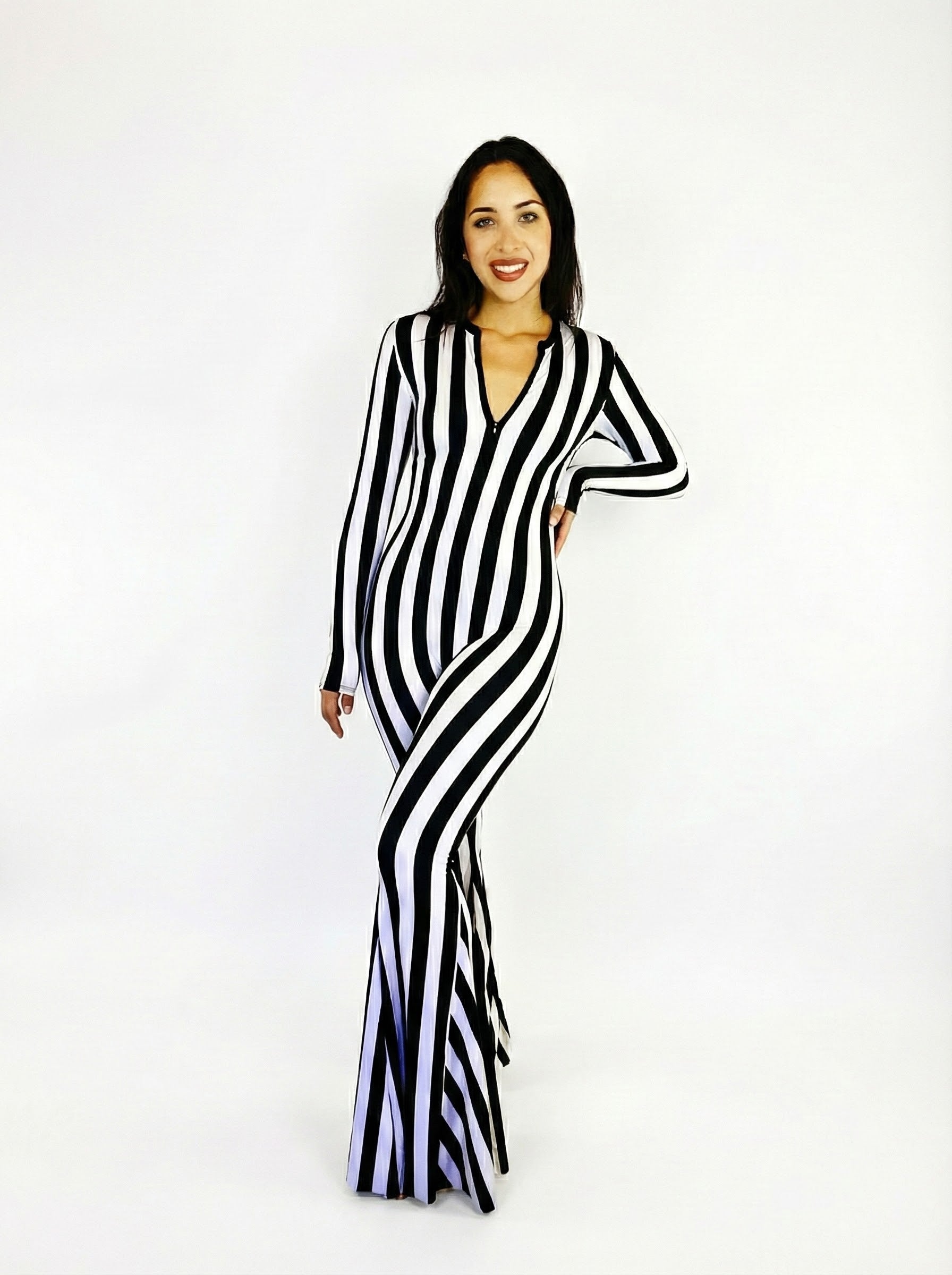 Model poses, smiling, in the Beetlejuice Black and White Striped Catsuit with bellbottom legs and a front zip, against a plain white background.