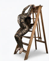 A contortionist in a Black Swirl On Holographic Catsuit with a scoop neck and full sleeves bends backward over a wooden stepladder.