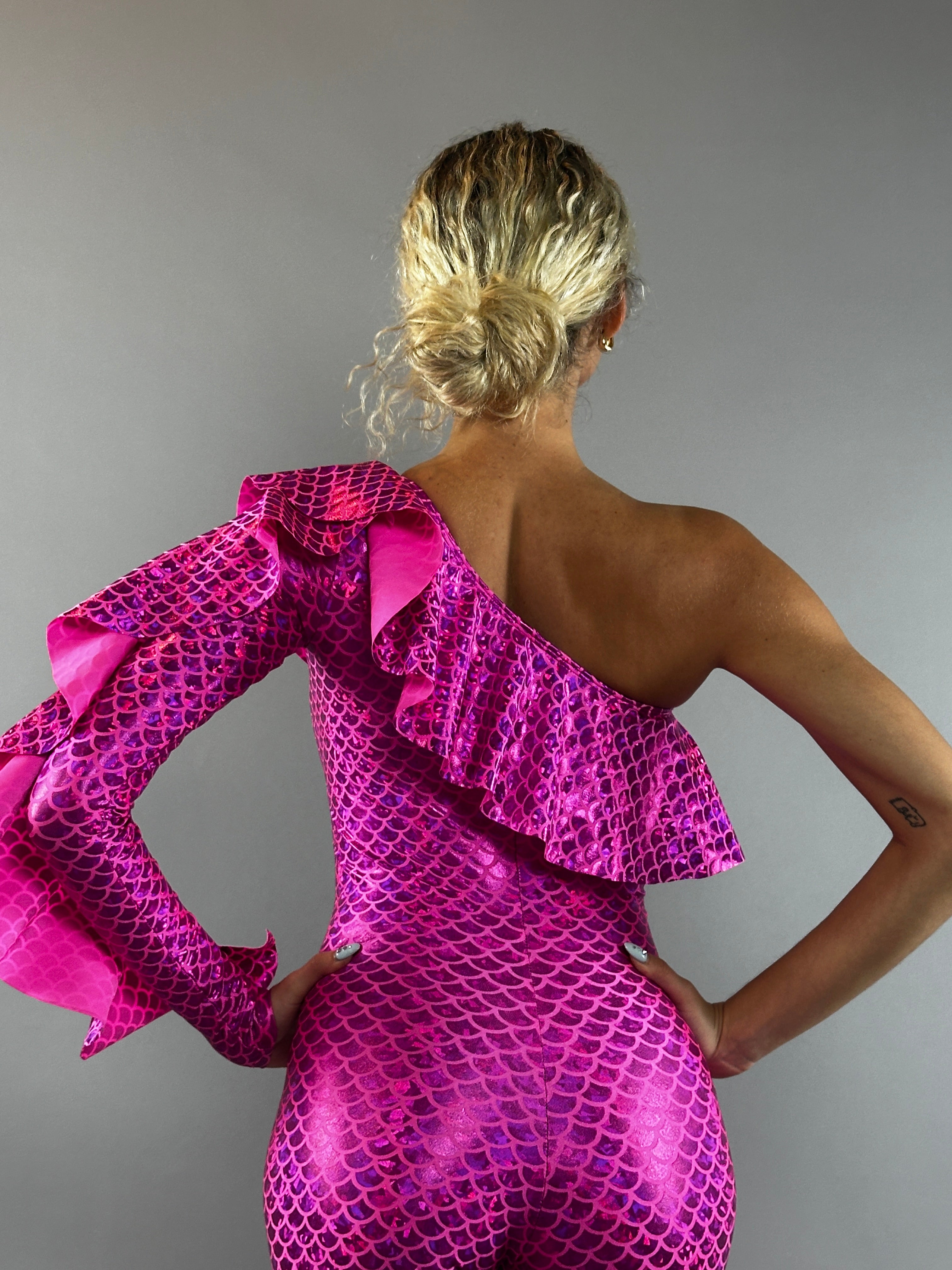 Person with blonde hair in a bun wears the Ruffle Catsuit, an off shoulder spandex jumpsuit featuring asymmetrical ruffles and a custom color scale pattern for a glamorous mermaid-inspired look.