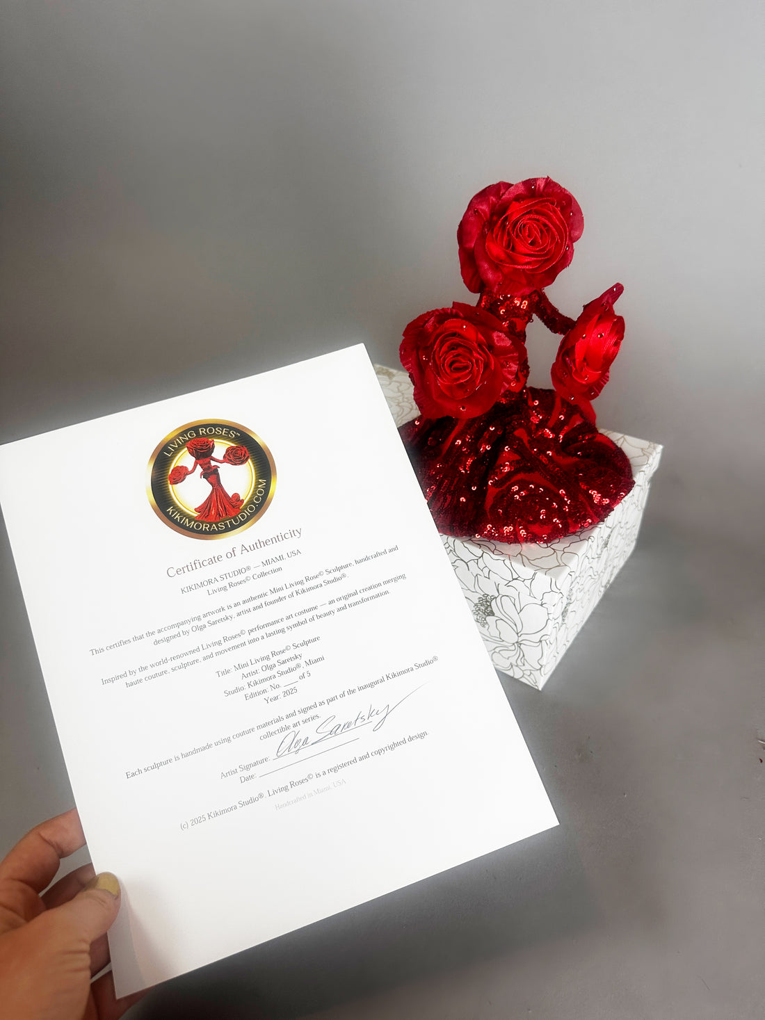 A hand holds a certificate beside a white box with three Mini Living Rose sculptures—doll ornaments by Kikimora Studio, each handcrafted and sparkling with red glitter.