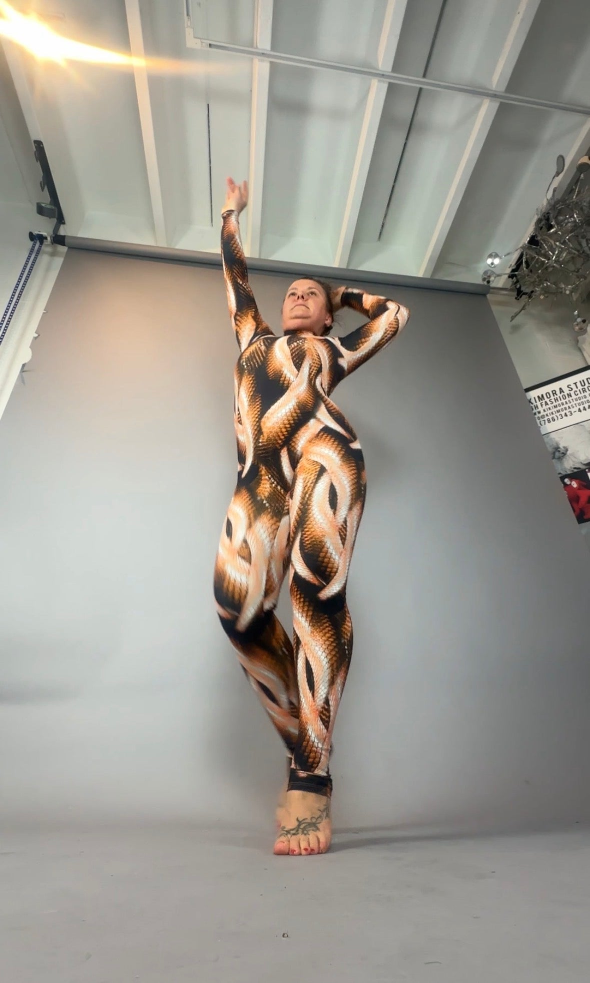 A person poses confidently in a full-body Brown Snake print Catsuit featuring a turtleneck and long sleeves, showcasing the bold metallic animal pattern against a plain backdrop.