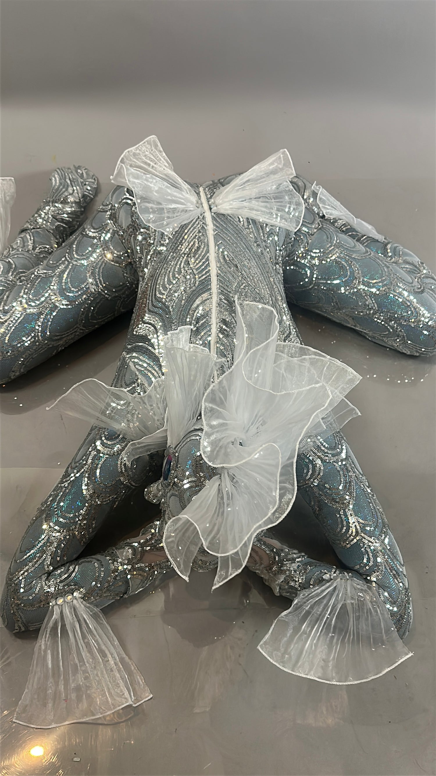 Sea Creature is a silver, textured frog plush adorned with white sheer ruffles on its head, neck, and limbs, giving it a couture-inspired look as it rests elegantly on a gray surface.
