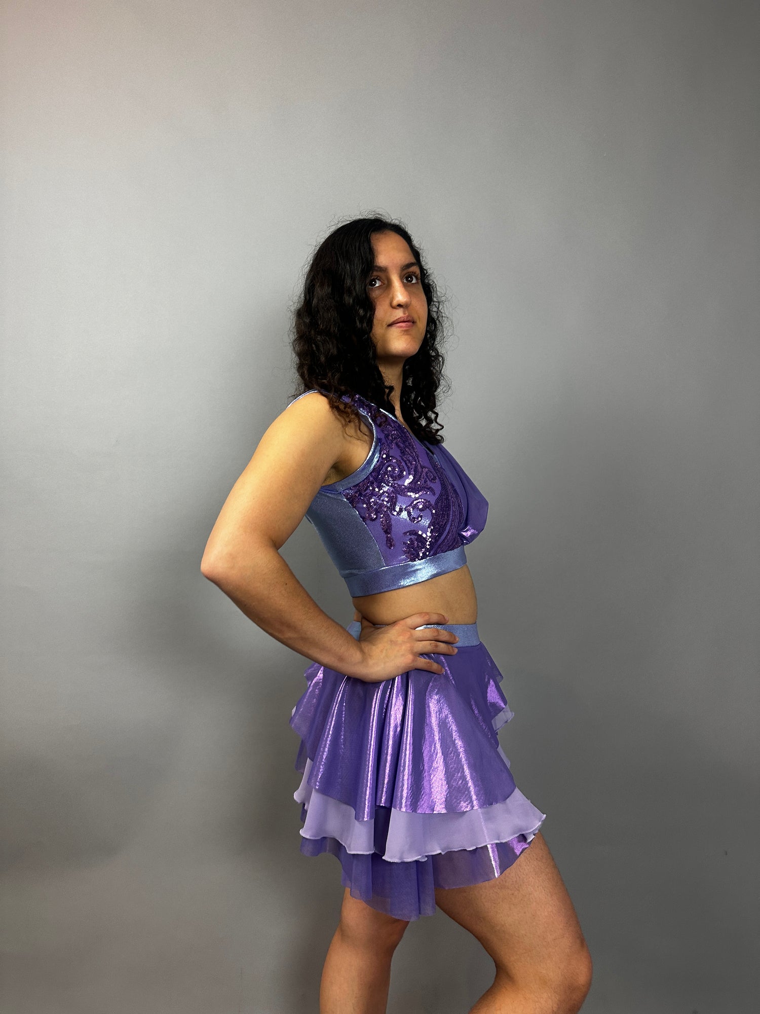A model poses confidently in the Lavender Purple Sequin Dance Costume—featuring a crop top, high waist shorts with half skirt, and 4-way stretch mesh—against a plain gray background.