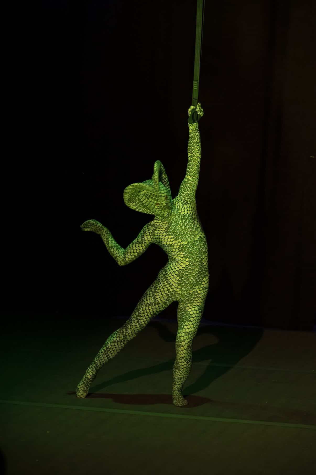 Model in a Green pixel Cobra Costume—Snake Print Catsuit with Serpent Headdress—strikes a dramatic pose on a dark stage, sword held overhead. This custom-made contortionist dance unitard is perfect for cosplay or performances.