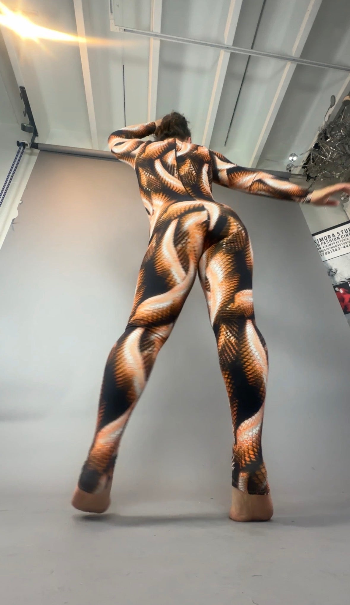 Person in a brown snake print catsuit with a turtleneck and long sleeves poses barefoot in a studio with a gray backdrop, facing away from the camera.