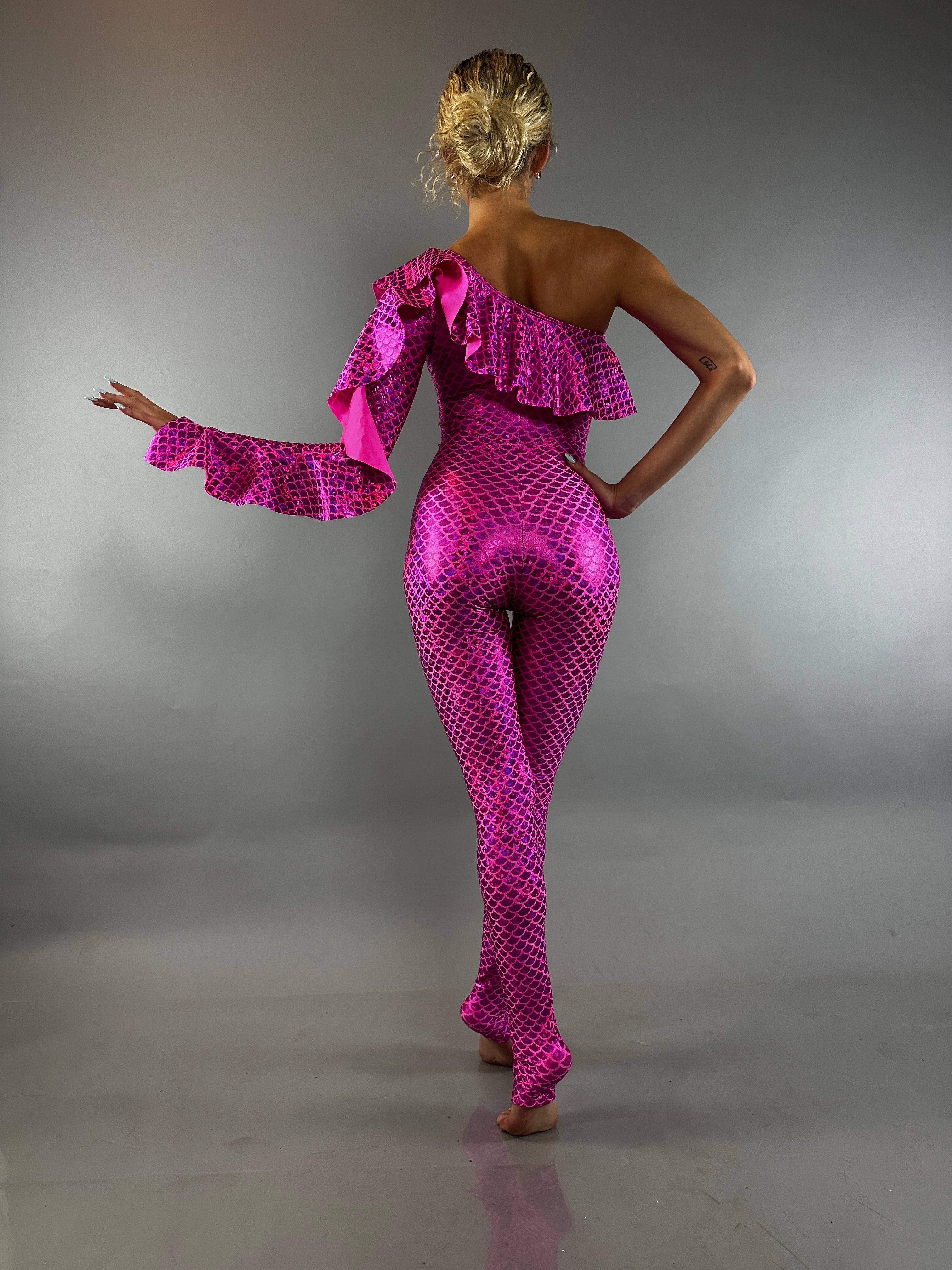 A woman wearing a Red Ruffle Catsuit—an off-shoulder, asymmetrical spandex jumpsuit in custom colors—stands barefoot against a gray background.