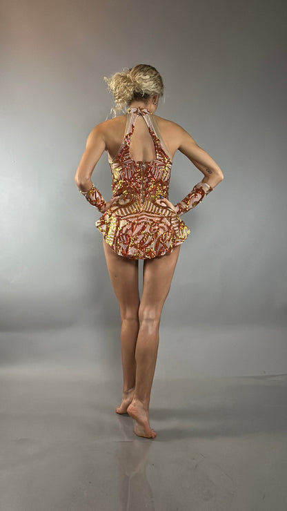 A woman stands barefoot with her back to the camera on a gray background, wearing the Fire Color Sparkling Halter Leotard, featuring red and gold sequin accents and an asymmetrical skirt that adds dynamic flair to her silhouette.