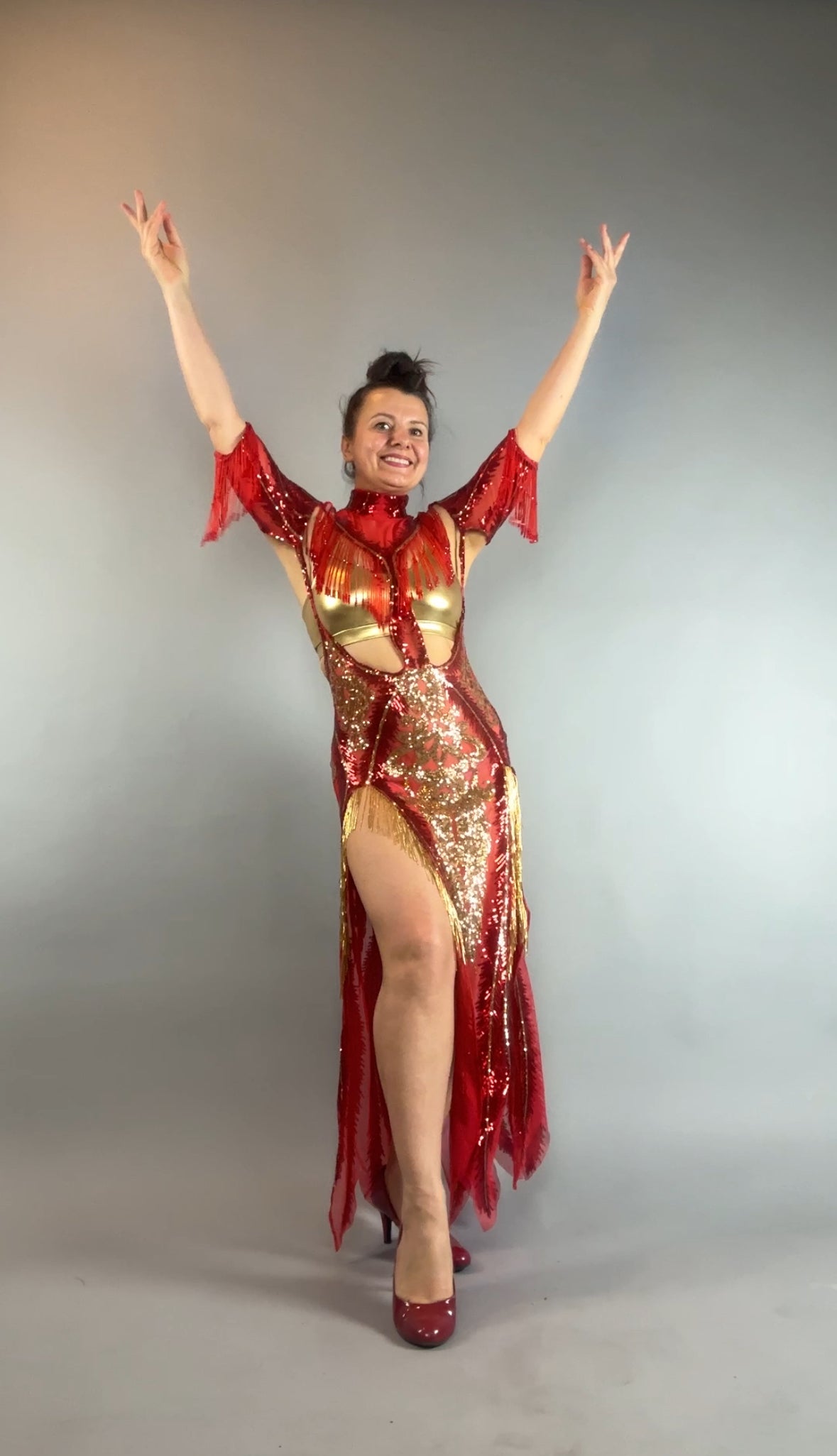 Woman wears the bearded French accessories Sequin Sheer Dress—backless with a thigh-high slit and beaded fringe—posing with arms raised and smiling against a plain background.
