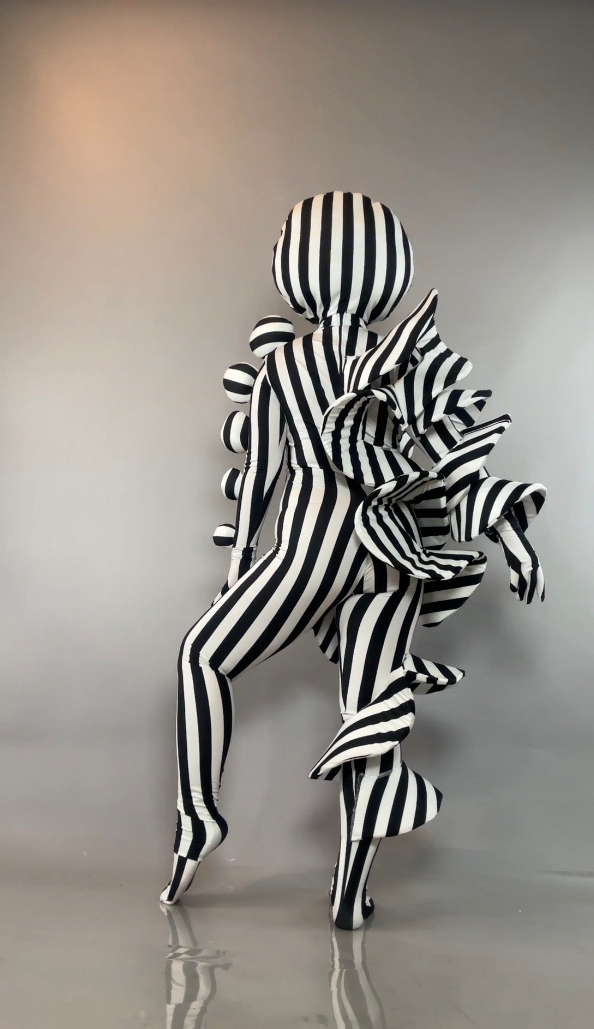 A person in a Black and White Striped Creature Costume with a spherical headpiece and sculptural ruffled back.