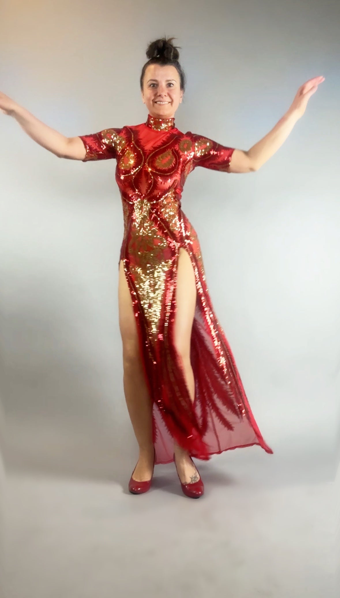 A person raises their arms against a plain background, wearing the Red And Gold Sequin Sheer Dress—sleeveless with dramatic high slit—and matching hand accessories.