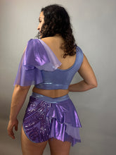 Woman models a Lavender Purple Sequin Dance Costume featuring high-waist shorts with a half skirt and crop top in 4-way stretch mesh, standing against a plain background, facing away.
