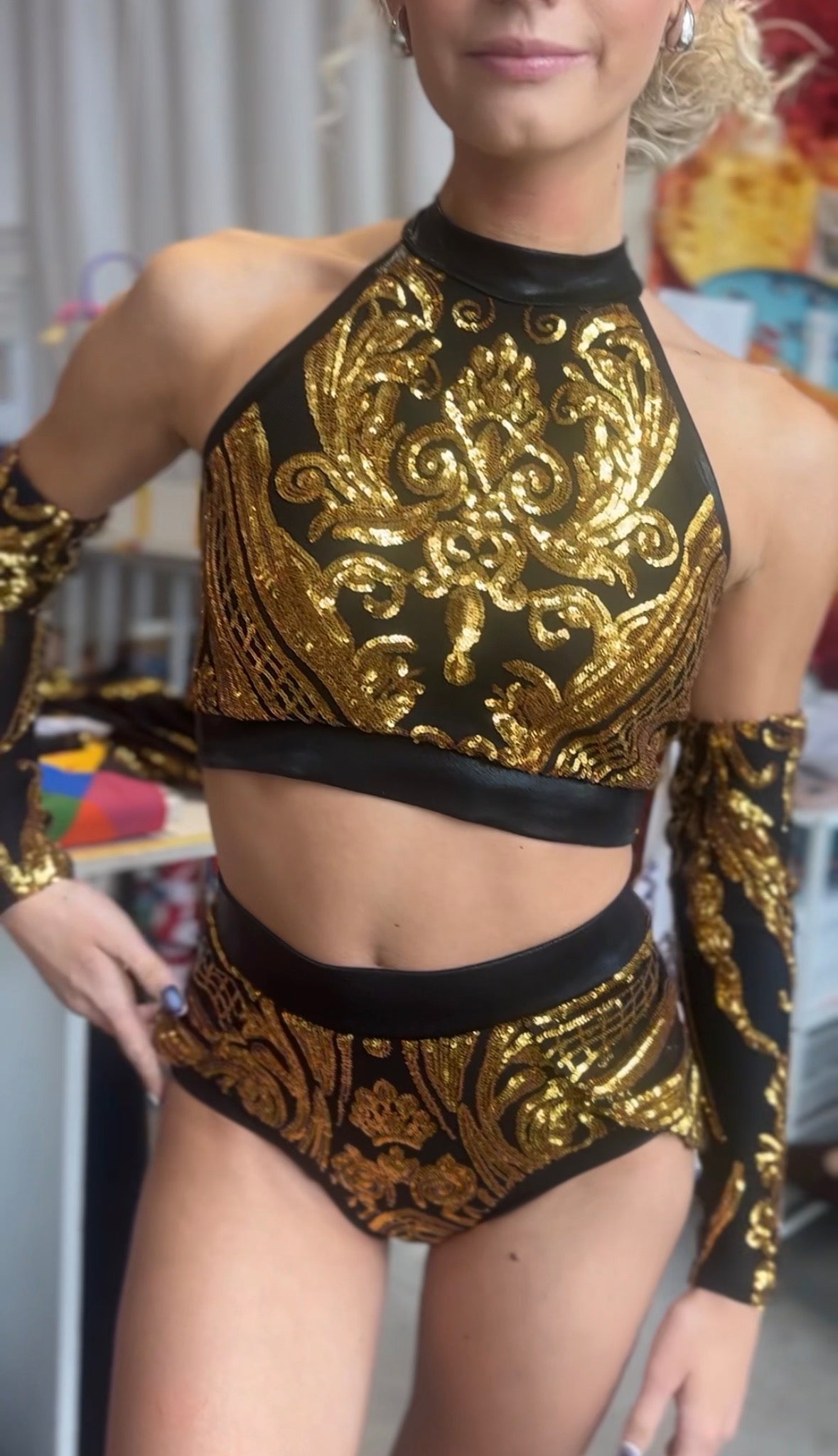 A woman wearing the Gold Sequin on Black Crop Top and Bottom Set, which features a halterneck crop top with ornate golden sequin detailing, matching high-waisted shorts, and elegant hand accessories.
