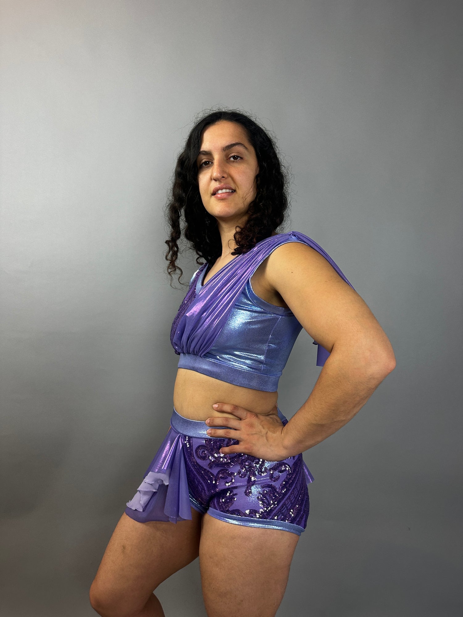 Wearing the Lavender Purple Sequin Dance Costume—featuring high waist shorts with a half skirt, crop top, and 4-way stretch mesh—the person smiles brightly against a plain gray background.