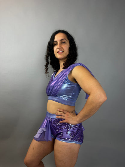 Wearing the Lavender Purple Sequin Dance Costume—featuring high waist shorts with a half skirt, crop top, and 4-way stretch mesh—the person smiles brightly against a plain gray background.