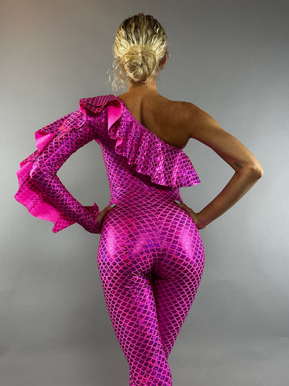A blonde-haired person in a bun wears the Ruffle Catsuit—an off-shoulder, asymmetrical spandex jumpsuit in shiny pink—posing against a gray backdrop.