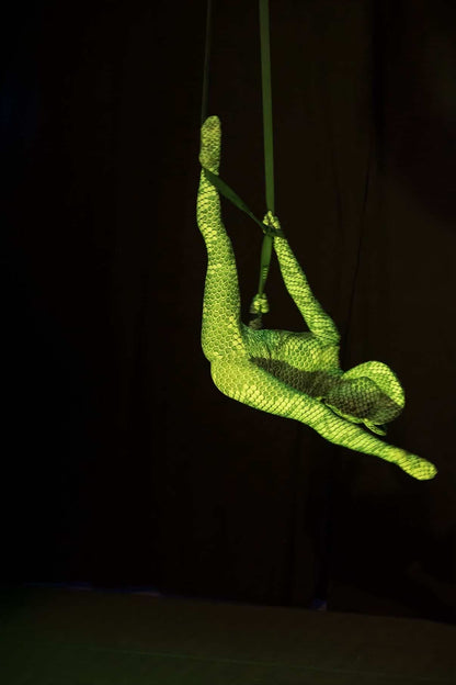An aerial performer hangs upside down on straps, her Green Pixel Cobra Costume— a snake print catsuit for dance and contortion—strikingly highlighted against a dark background.