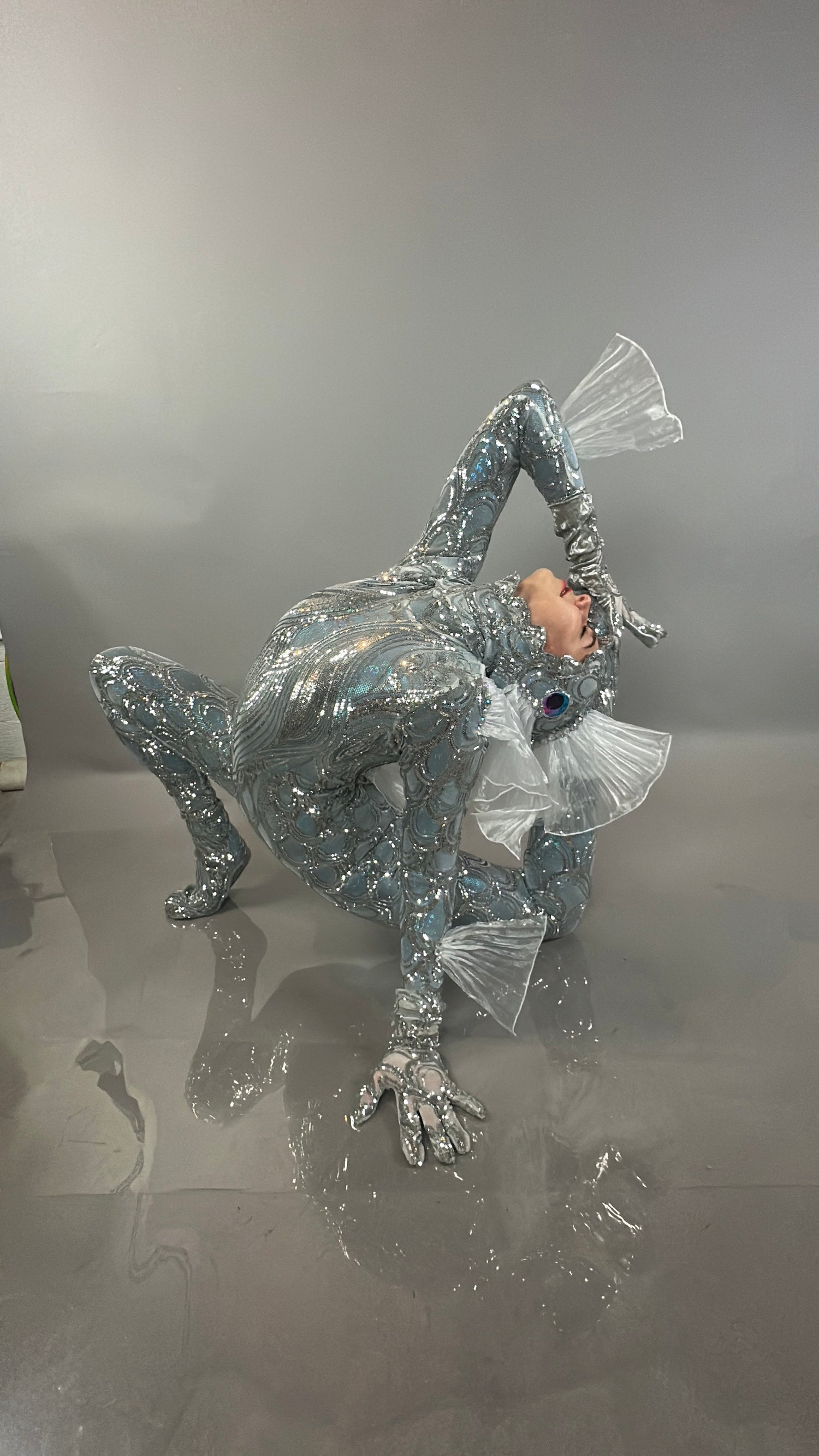 A person models the Sea Creature, a shiny silver costume with fins, striking a dramatic pose on a glossy gray surface with a matching backdrop—showcasing wearable sculpture and couture design.