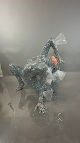 A person models the Sea Creature, a shiny silver costume with fins, striking a dramatic pose on a glossy gray surface with a matching backdrop—showcasing wearable sculpture and couture design.
