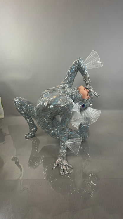 A person models the Sea Creature, a shiny silver costume with fins, striking a dramatic pose on a glossy gray surface with a matching backdrop—showcasing wearable sculpture and couture design.