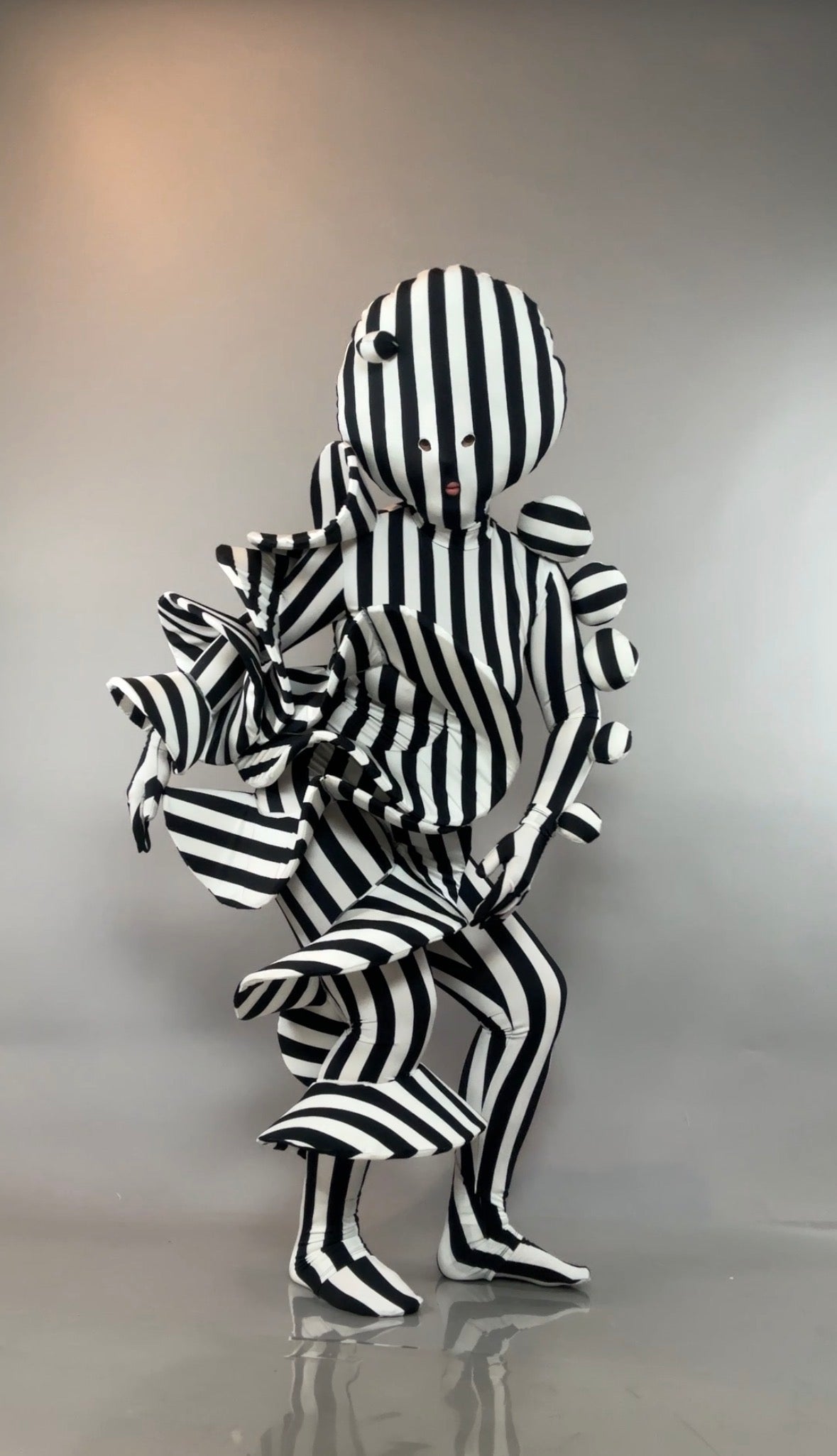 A person in a Black and White Striped Creature Costume with sculptural, ruffled elements, a mask, and matching gloves.