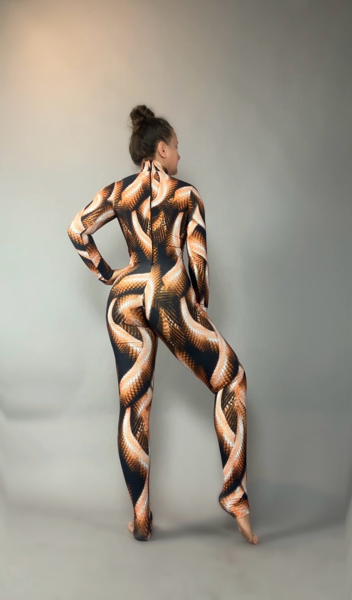 A woman poses barefoot against a plain gray background, facing away, wearing the Brown Snake Print Catsuit with a turtleneck and long sleeves.
