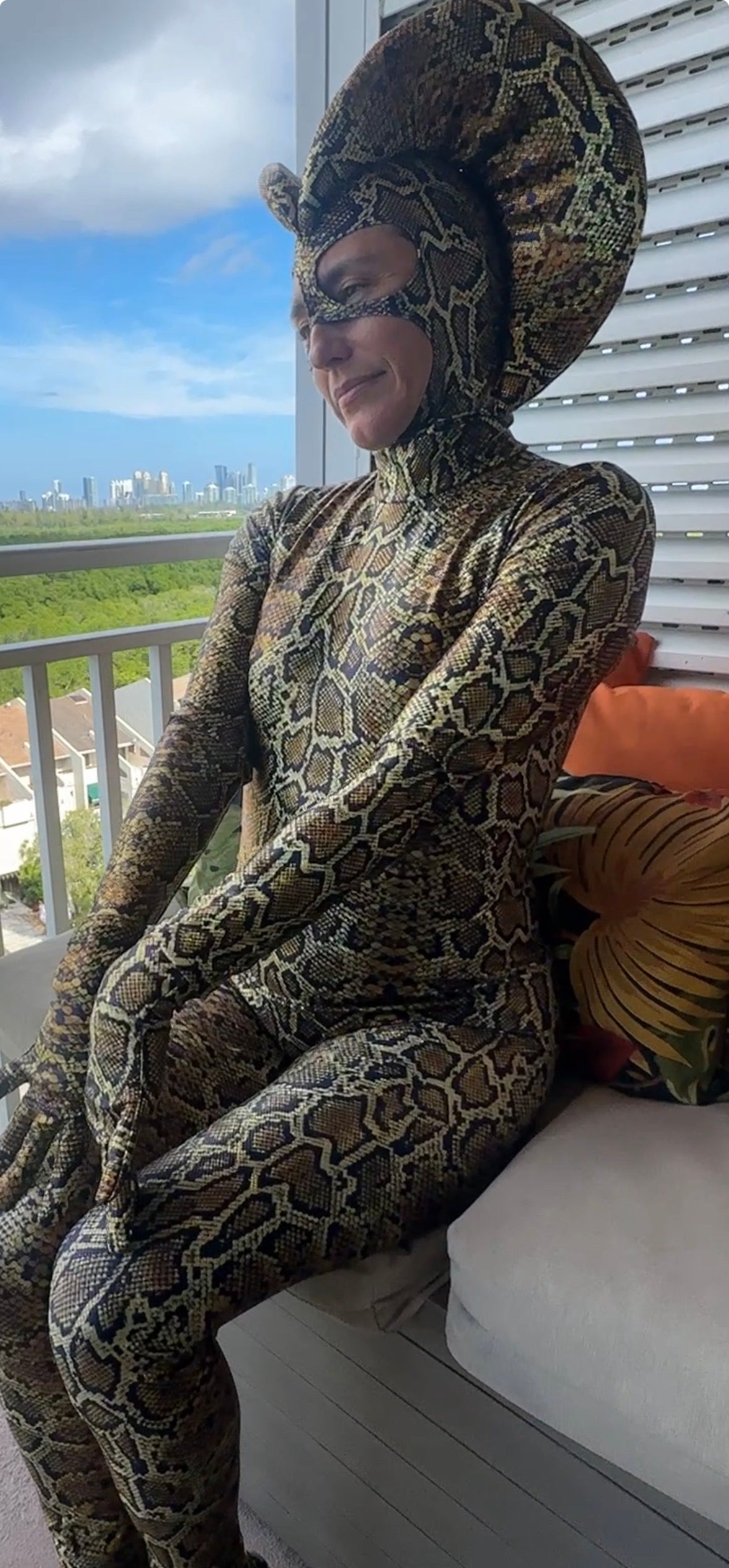 A person in a Green Cobra Snake Costume—ideal for gymnastics, acrobatics, dancers, and contortionists—sits on a balcony with city and greenery in the background.