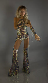 A woman wearing a sequined Chaps pants and laced top festival costume set poses in tall platform boots, adjusting her sleeve against a plain gray background.