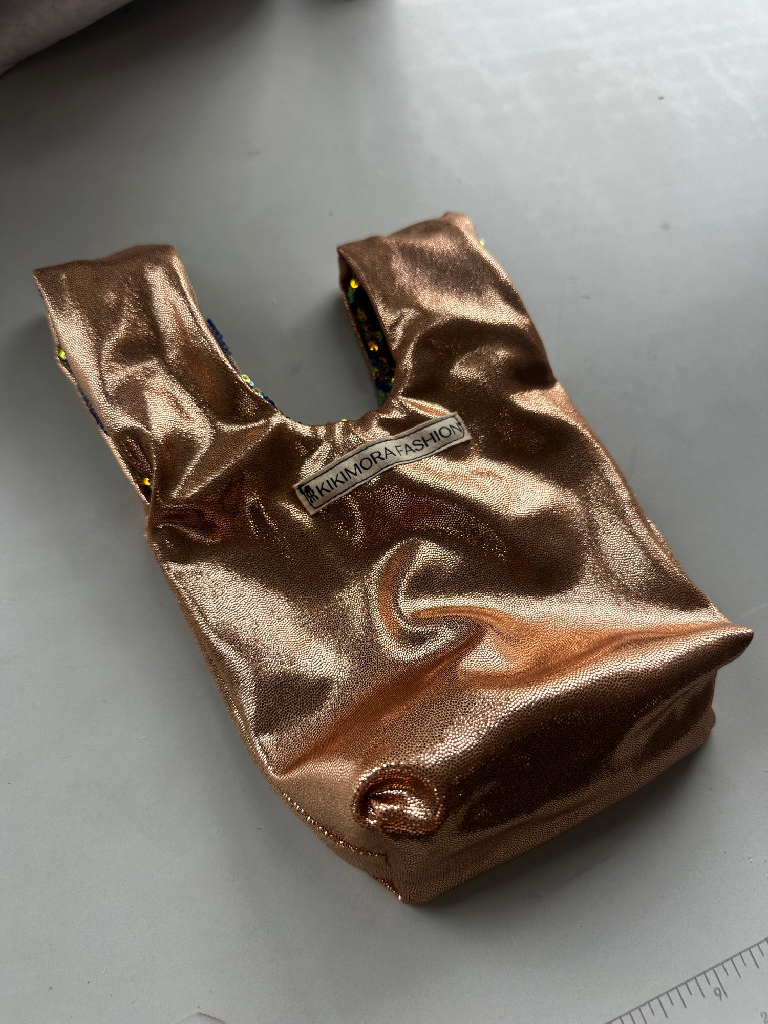 The handcrafted reversible sequin mini tote, a sustainable fashion accessory, features shiny bronze sequins and brings glam to any outfit; labeled “KIKIMORE FASHION” and shown on a light gray surface.