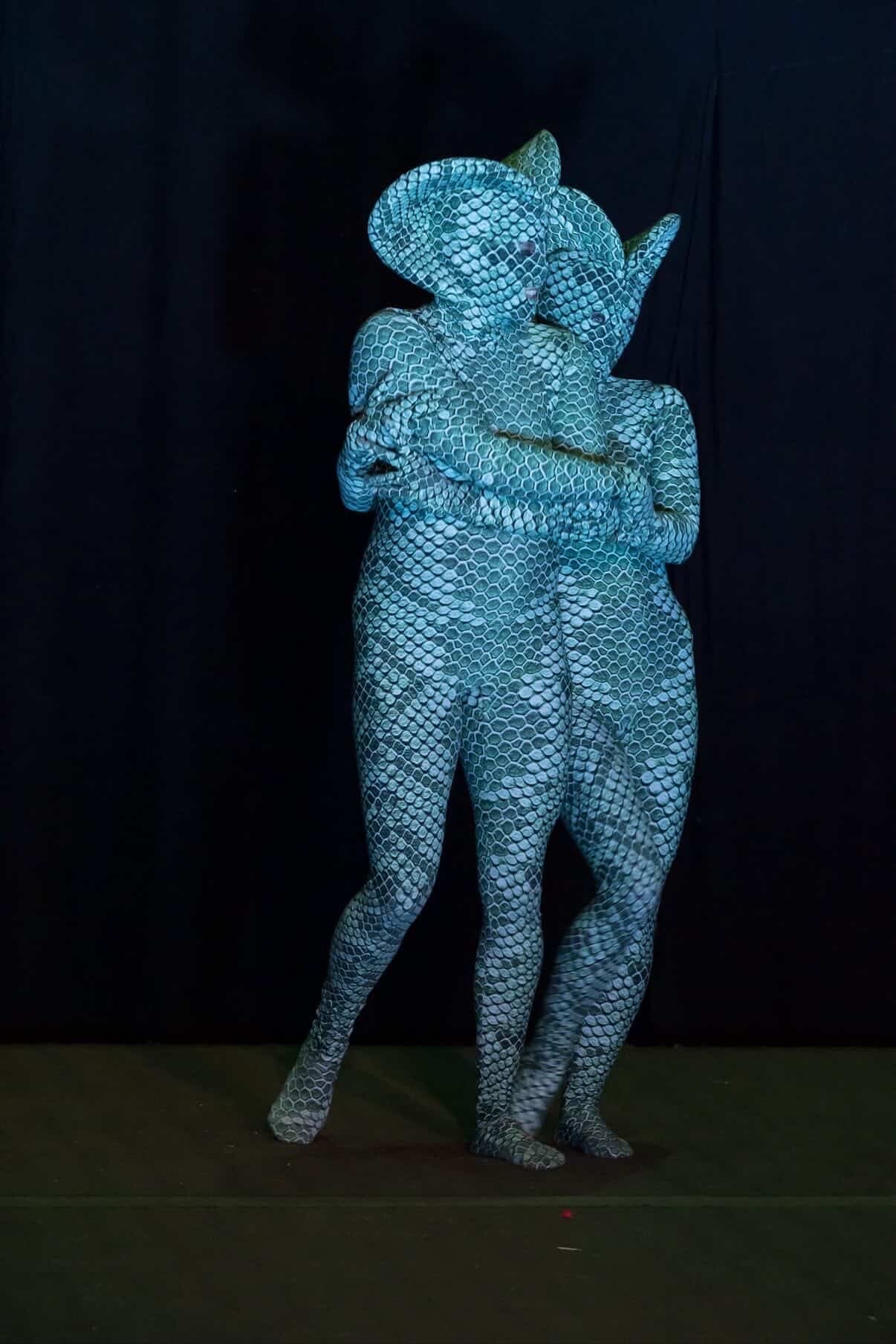 Two people wearing Green pixel Cobra Costumes—snake print catsuits with horned headdresses—embrace against a dark background. This custom-made serpent cosplay is ideal as a contortionist outfit, dance unitard, or unique performance piece.