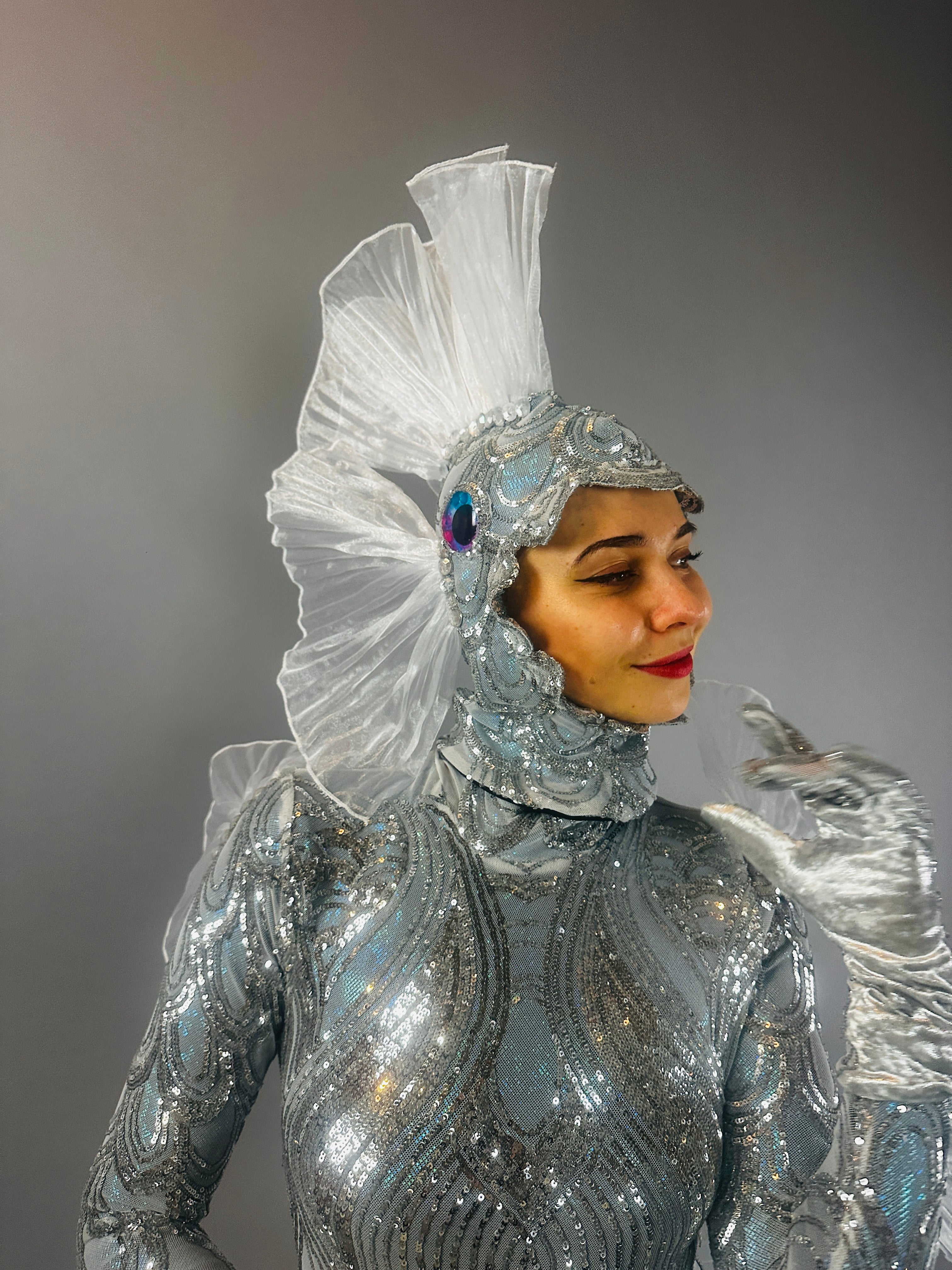 Model wears "Sea Creature": a shiny silver, fish-inspired couture costume with a fin-like headpiece, resembling wearable sculpture against a gray background.