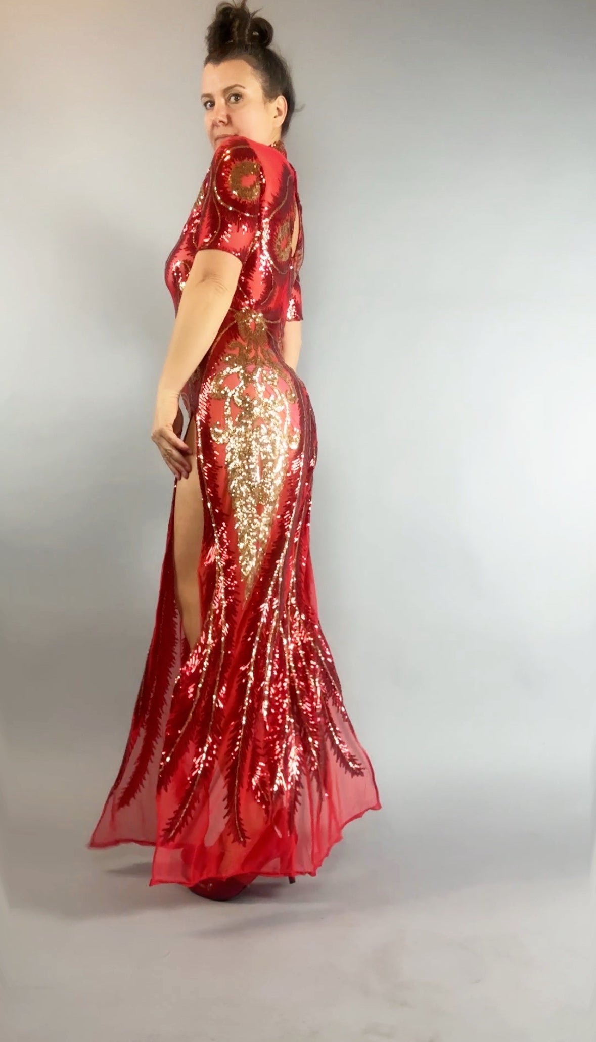 Woman in a Red And Gold Sequin Sheer Dress, sleeveless, styled with hand accessories, poses elegantly against a plain gray background.