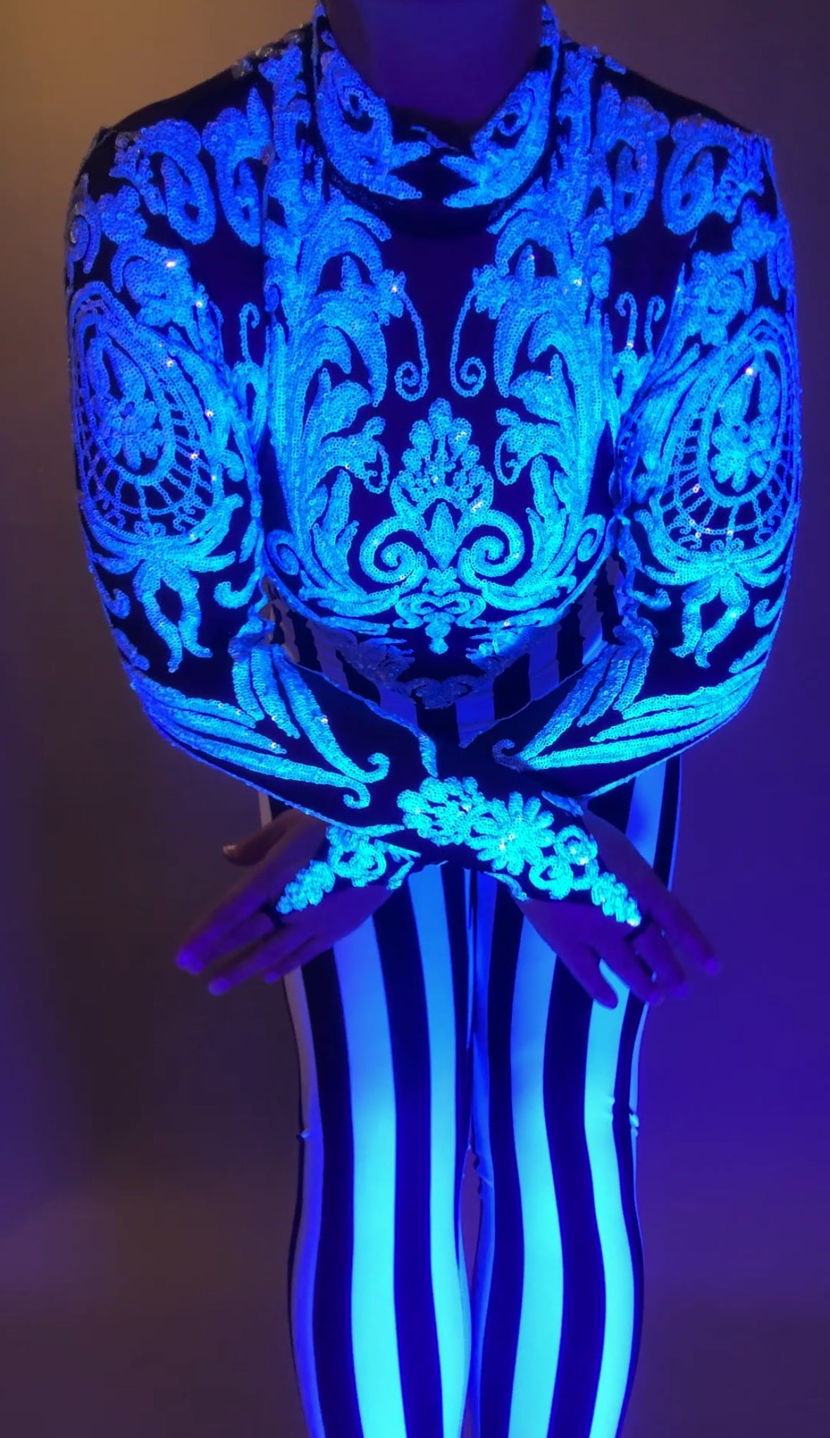 Wearing the Glamorous sequins Stripes Catsuit costume, a person stands with arms crossed, in vivid lighting. The blue and white striped outfit glows with ornate detail.