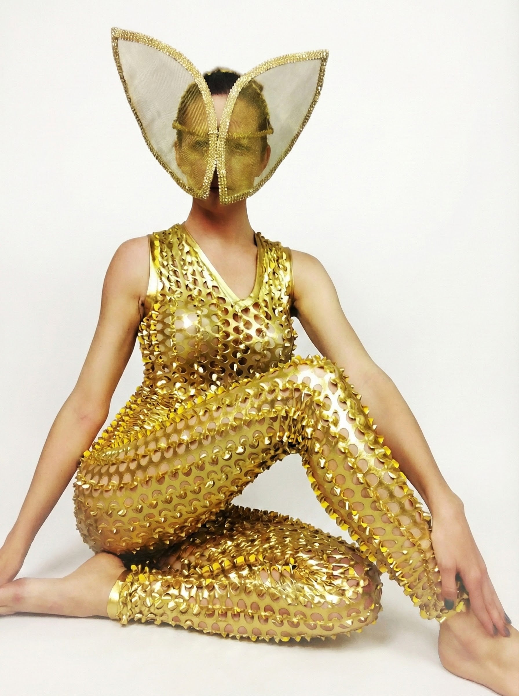 Person in a Gold Hollow Out Catsuit with a scoop neckline and sleeveless design, featuring cutouts and a large gold butterfly-shaped face mask, sitting cross-legged.
