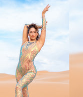 A woman wearing an Iridescent Sequin Skintone Catsuit with a halterneck and sleeveless design poses with arms raised in the desert beneath a blue sky.