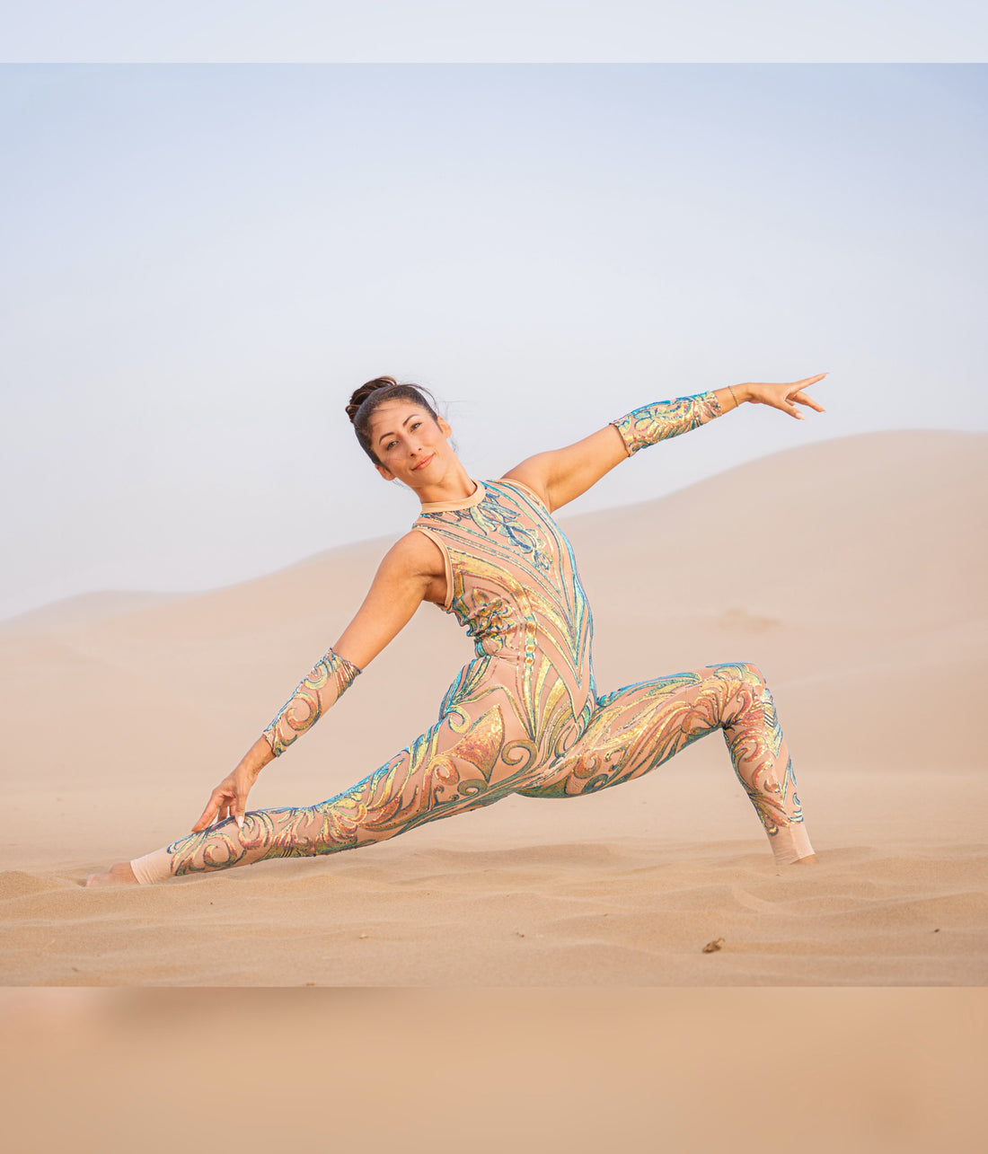 A woman wearing the Iridescent Sequin Skintone Catsuit with a halterneck and sleeveless design poses in a split on sand dunes under a clear sky.