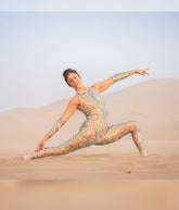 A woman wearing the Iridescent Sequin Skintone Catsuit with a halterneck and sleeveless design poses in a split on sand dunes under a clear sky.
