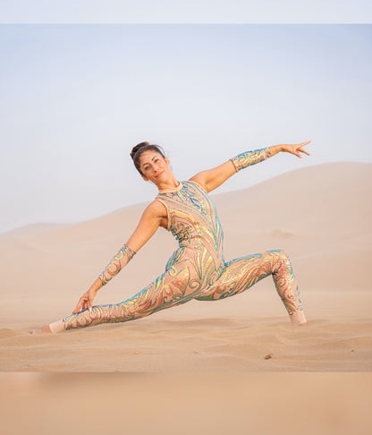 A woman wearing the Iridescent Sequin Skintone Catsuit with a halterneck and sleeveless design poses in a split on sand dunes under a clear sky.