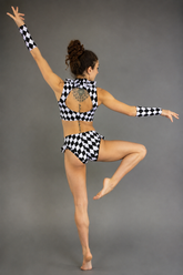 Wearing the Ruffled Harlequin Dance Costume—a black and white checkered spandex set with high waist shorts, a crop top, and matching arm sleeves—a dancer poses on one leg with arms extended, shown from the back.