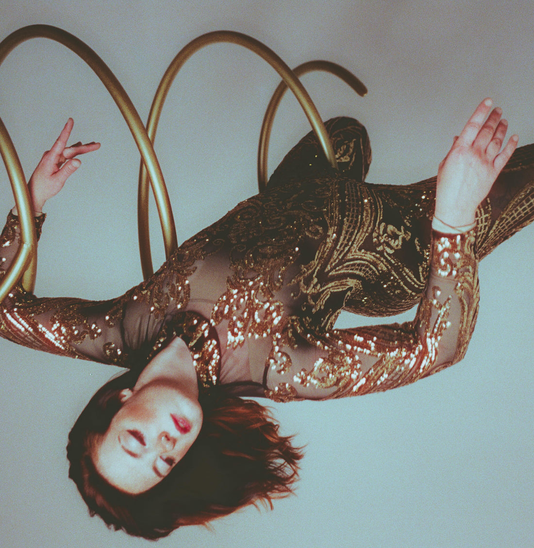 A person in a Black and Gold Sequin Catsuit with full sleeves and a turtleneck hangs upside down within golden spirals, eyes closed, illuminated by dramatic lighting.